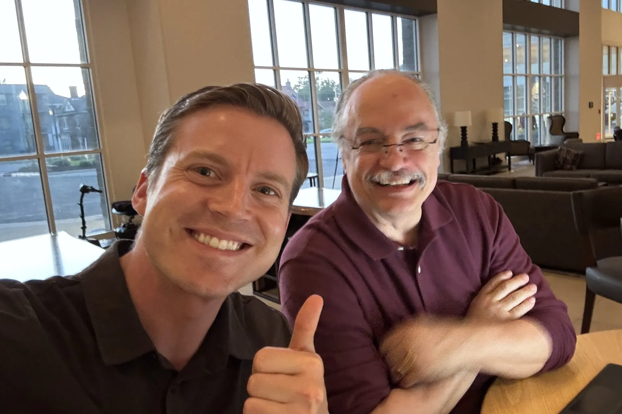 Stefan Bauer giving a thumbs up in a selfie with a laughing Dr. Ortiz at a table in a hotel lobby.