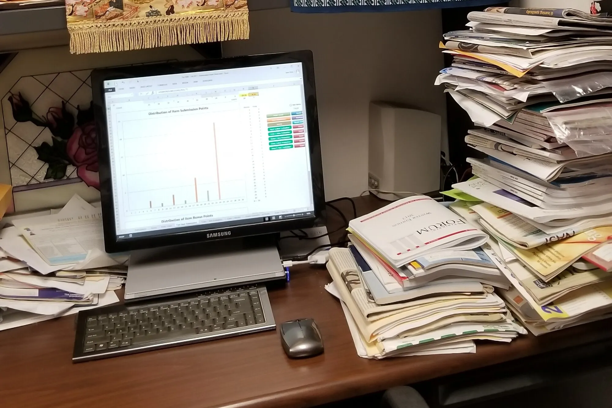 A desk with a computer monitor showing a chart, surrounded by tall stacks of papers and journals.